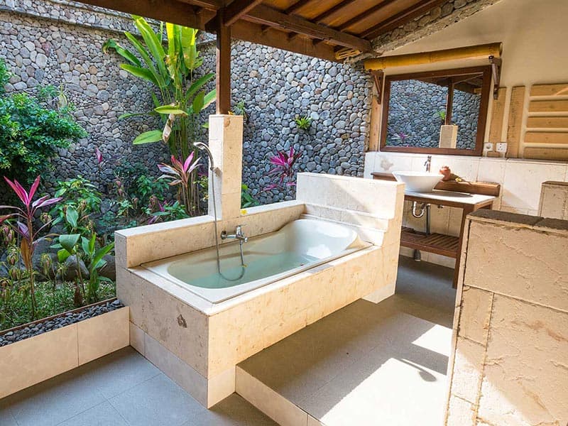 Bathroom with sunken soaking tub, twin sinks, wooden beams, stone accent wall, and tropical plants visible through open areas
