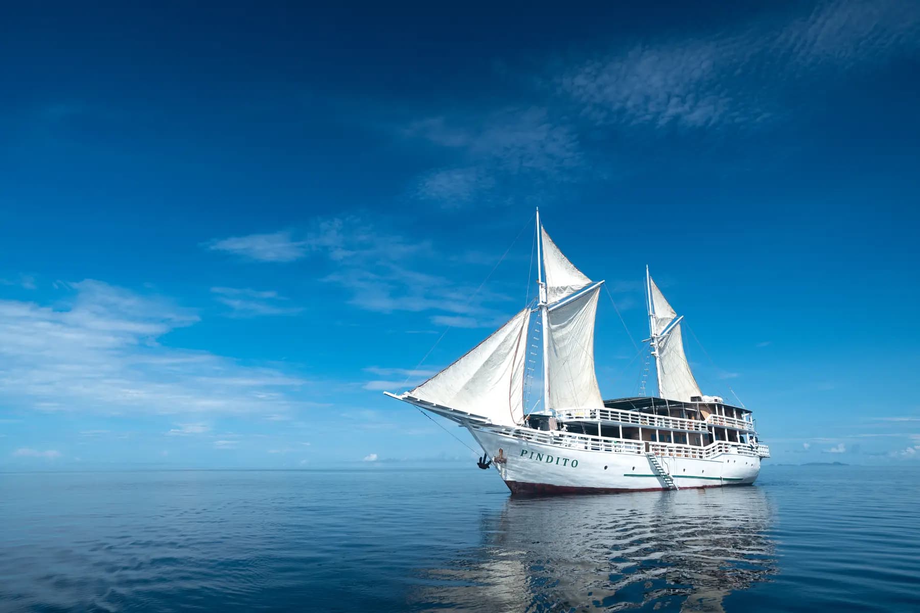 The Pindito liveaboard with white sails raised, anchored on calm blue water under a clear sky