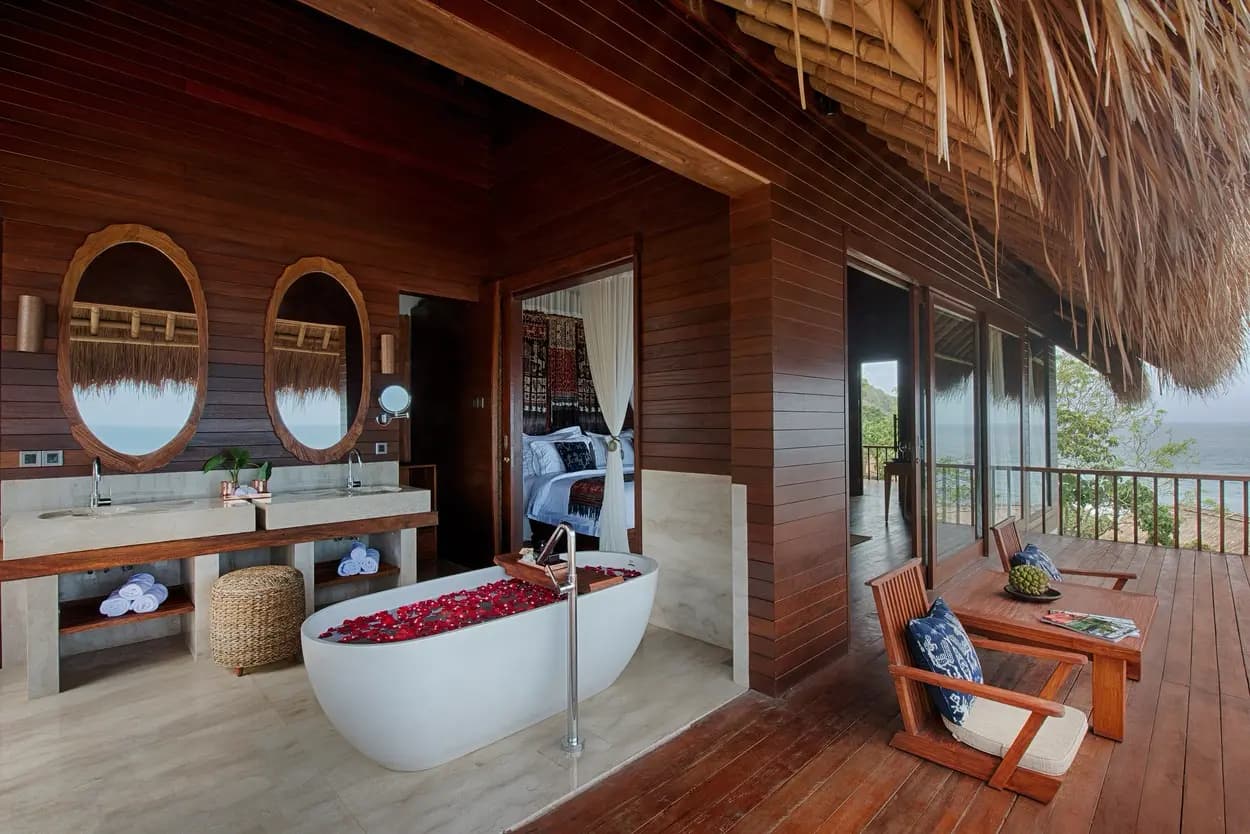 Bathroom with dual sinks, oval mirrors, white freestanding tub filled with red petals, and an open deck with wooden seating and thatched roof structure