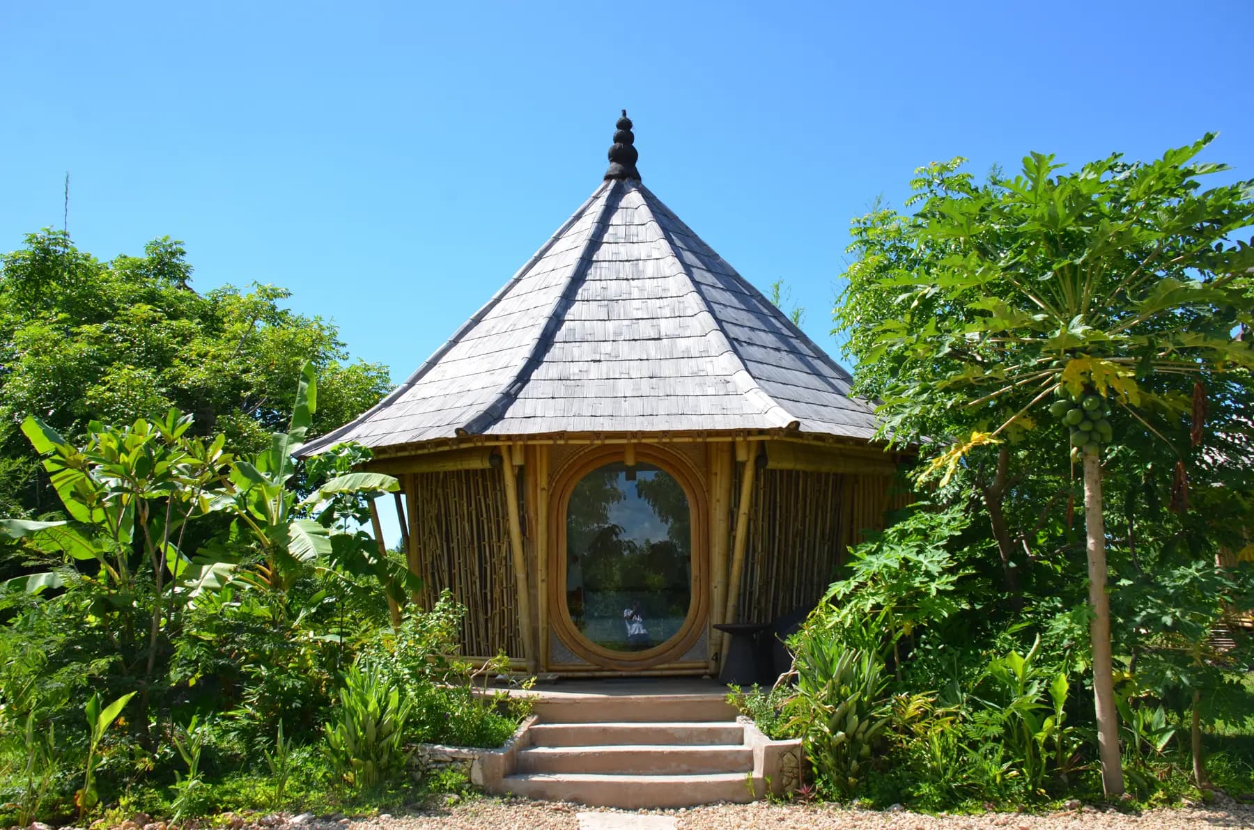 A conical wooden structure with a slate roof and large oval window, surrounded by lush tropical vegetation and stone steps leading to its entrance
