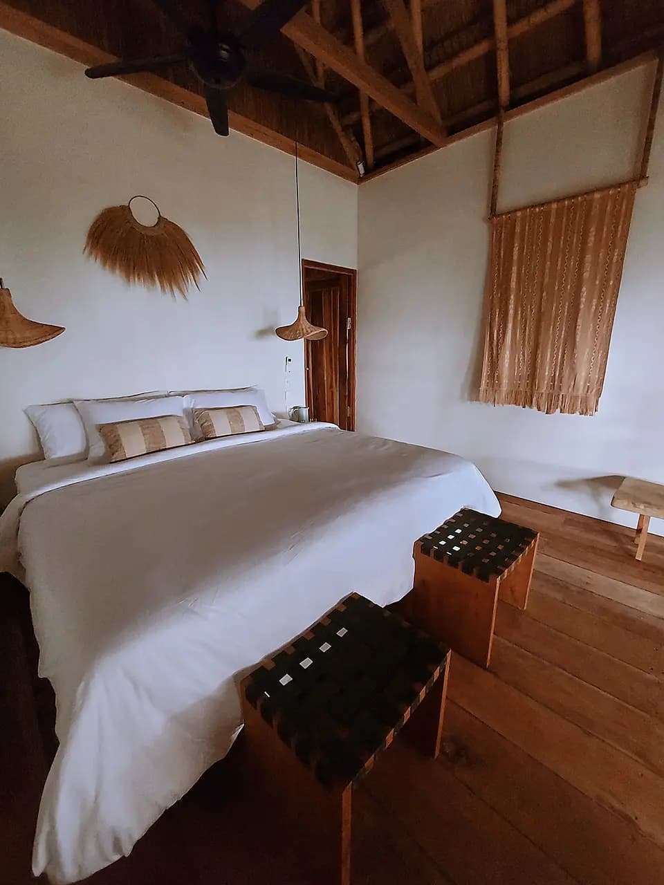 Bedroom with white bedding, woven pendant lights, wooden ceiling beams, striped cushions, and wooden flooring