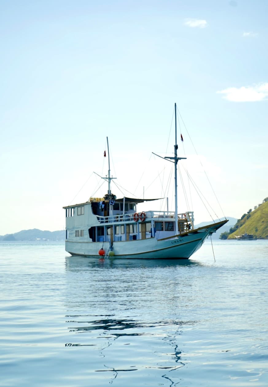 The liveaboard Umami anchored on calm water with two masts raised, hills visible in the distance