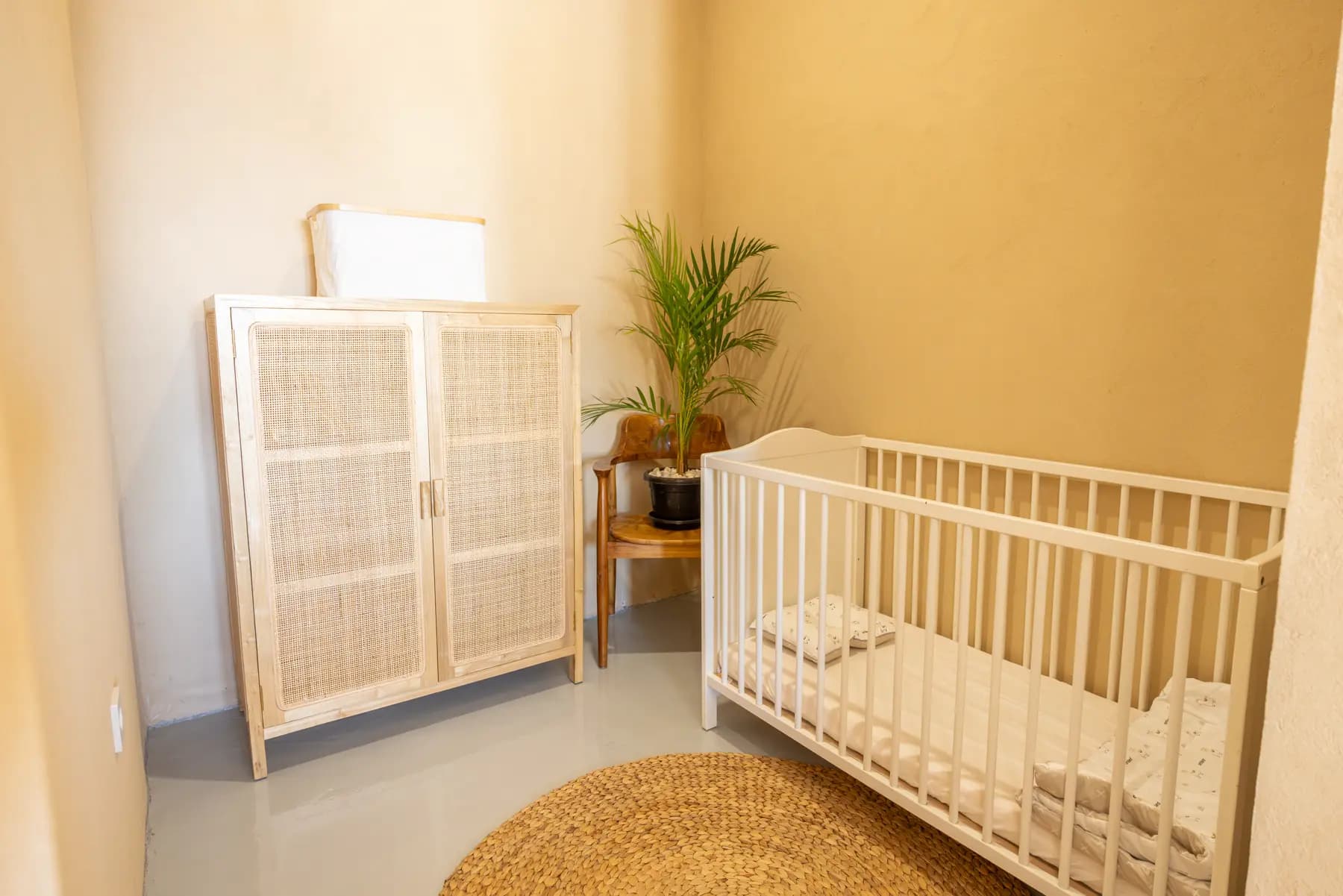 Nursery room at Jepun Hill Studios with a white crib, rattan cabinet, potted plant, and woven floor mat
