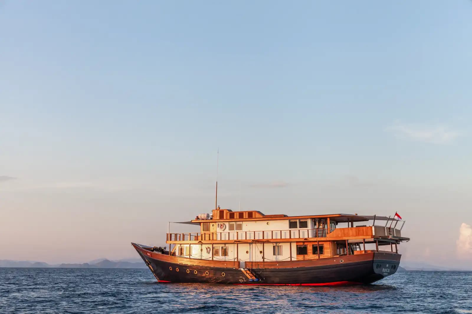 The Rebel liveaboard anchored on calm water at dusk, with distant hills faintly visible on the horizon