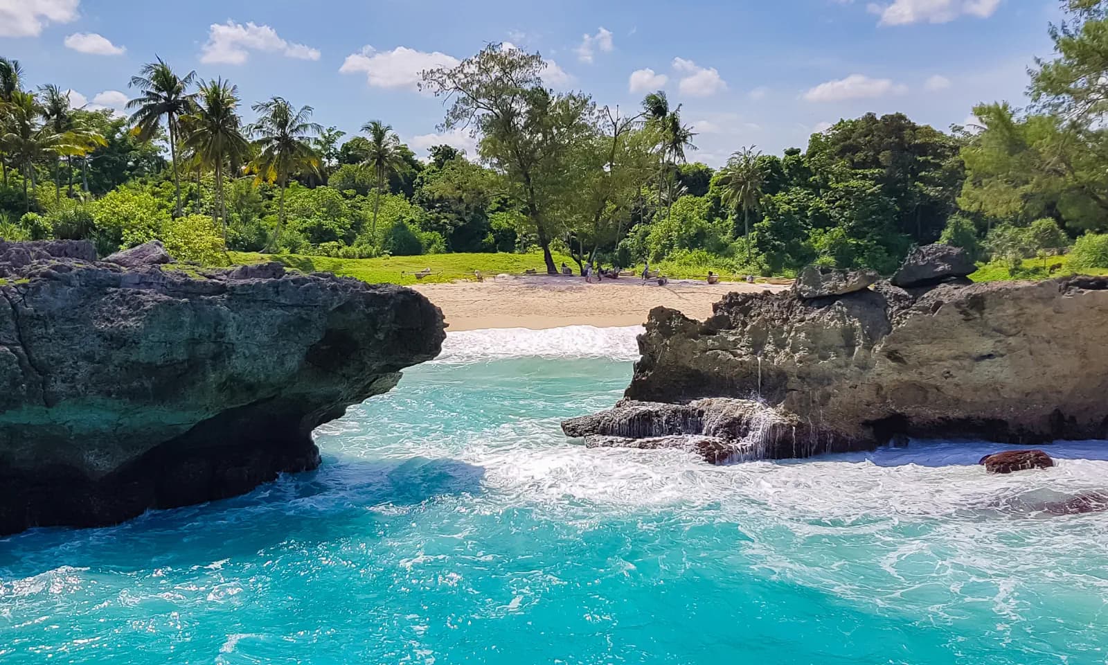 Turquoise water flowing between dark rock formations toward a sandy beach lined with palm trees and greenery