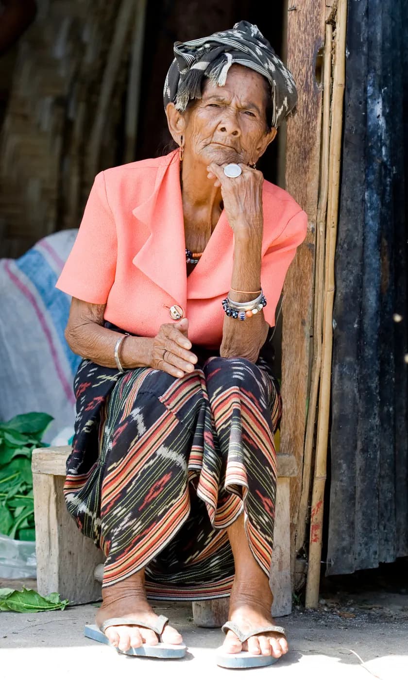 Elderly woman in a pink jacket and patterned sarong sits on a wooden bench wearing a headscarf and bracelets