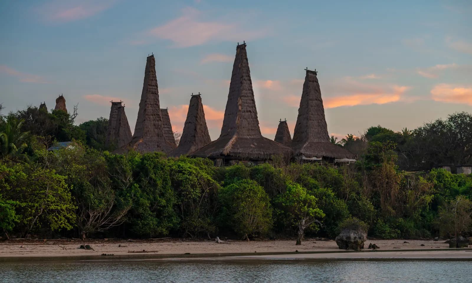 Tall thatched-roof peaked houses rising above dense trees along a shoreline at dusk with a pink-streaked sky