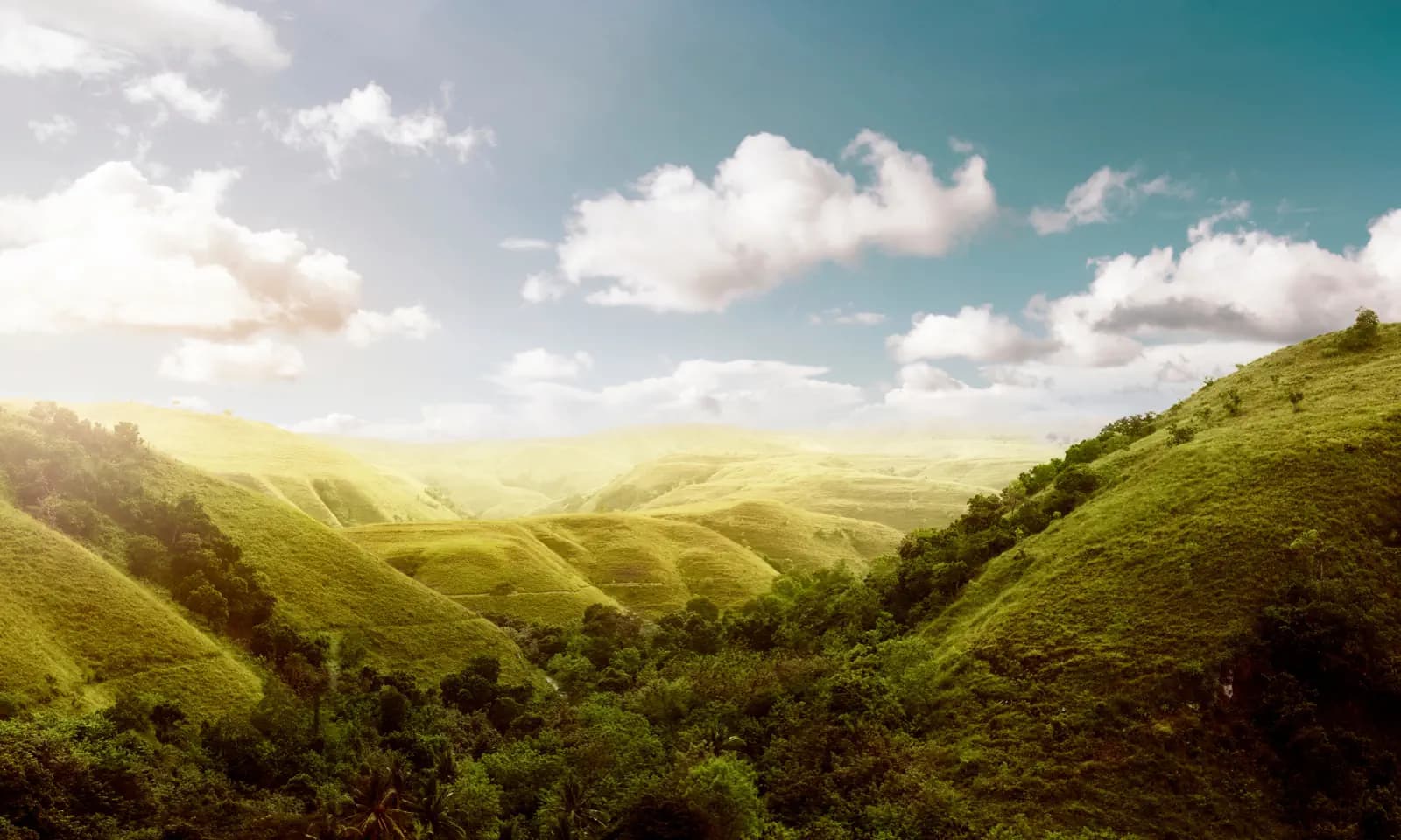 Rolling green hills and a forested valley under a blue sky with scattered white clouds and hazy sunlight