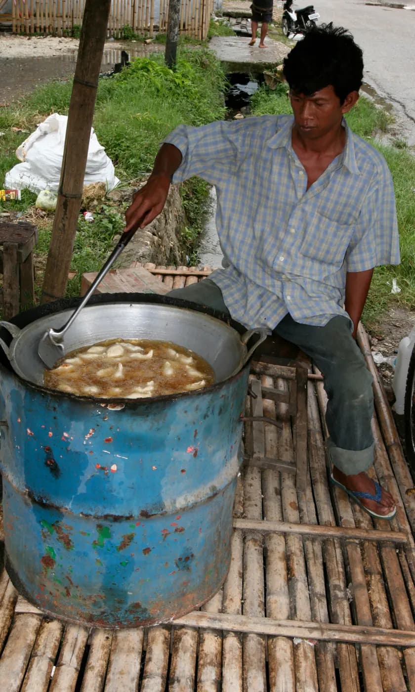 Man sitting beside a large blue drum, stirring food frying in oil with a long metal ladle
