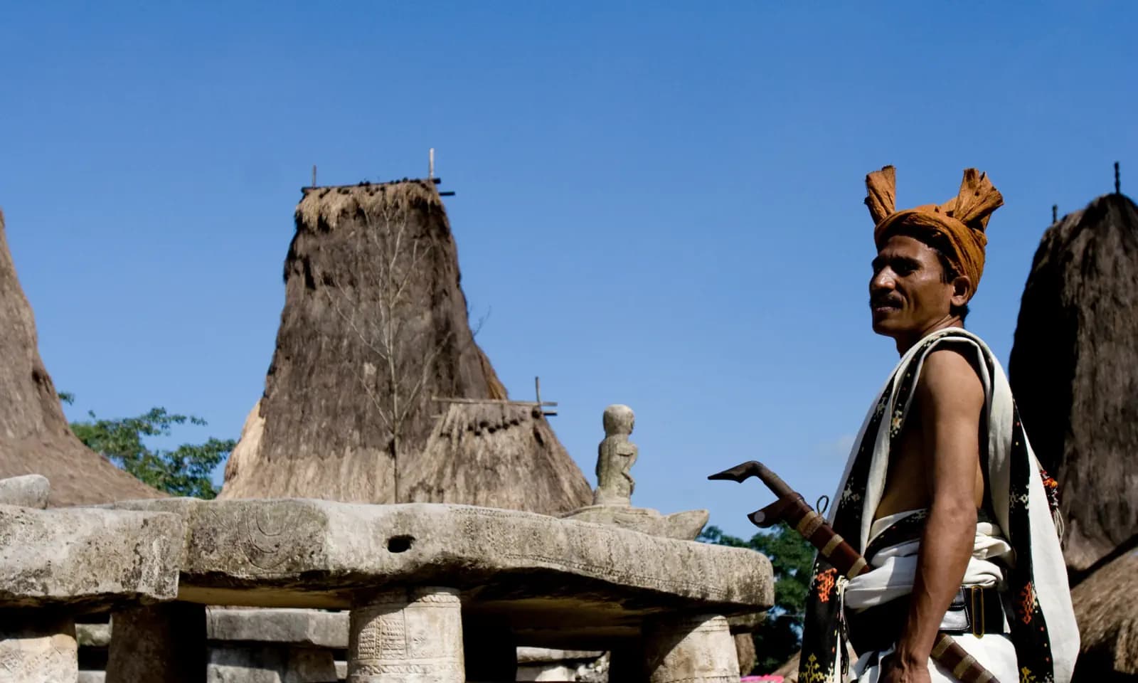 Man in a traditional headwrap and carved knife standing near stone tombs and thatched-roof houses