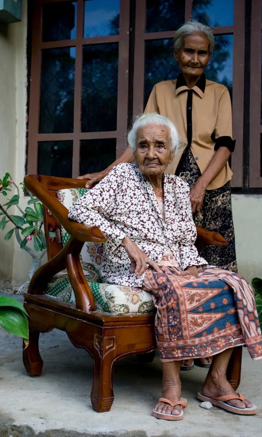 Elderly woman seated in a wooden armchair with a batik cloth on her lap, another woman standing behind her