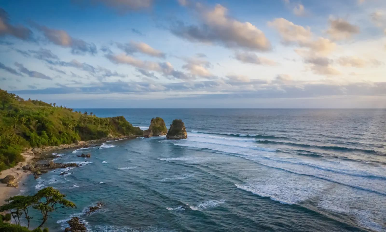 Rocky sea stacks rise from waves near a green coastline, with a cloudy sky at sunset