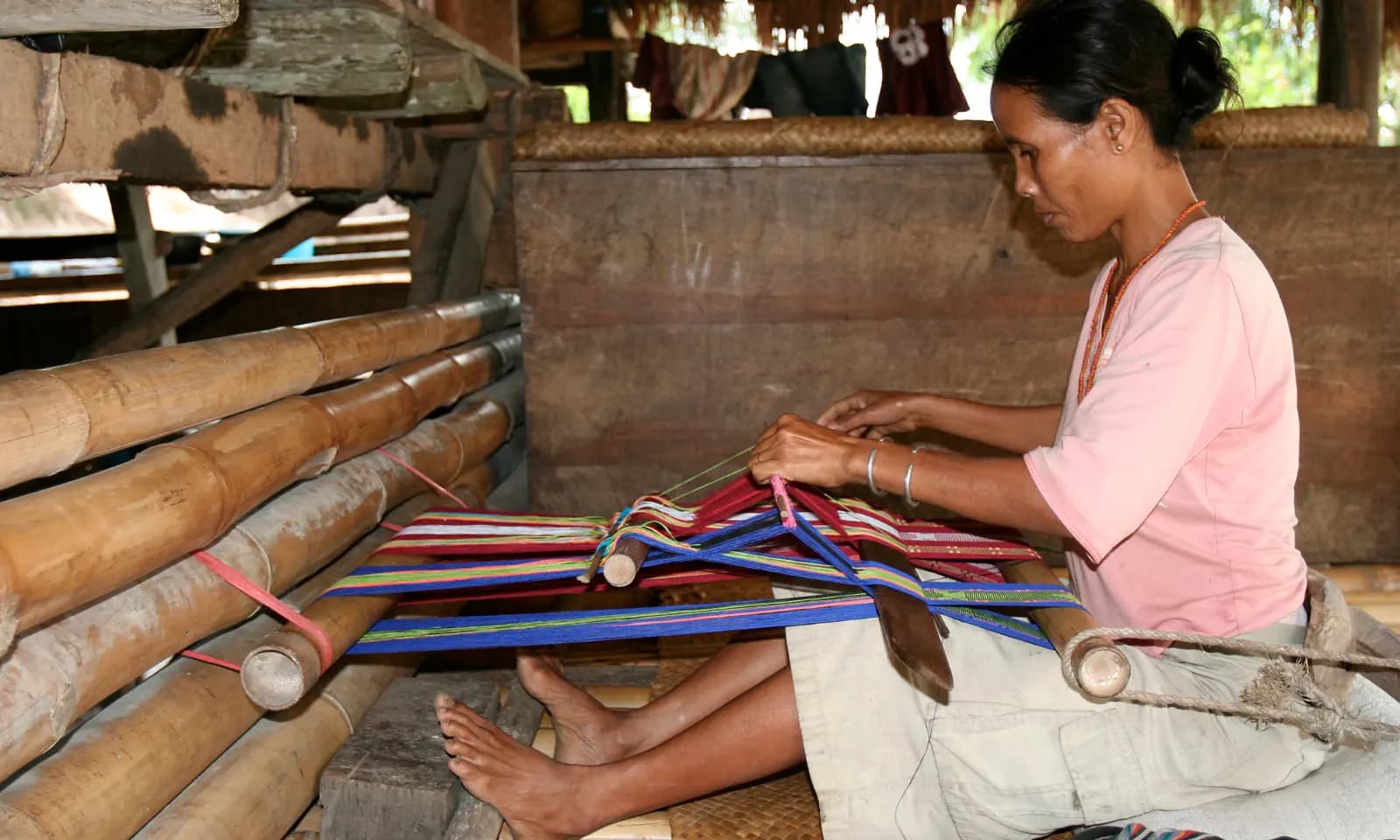 Woman sitting on a floor mat weaving colorful threads on a backstrap loom under a wooden shelter