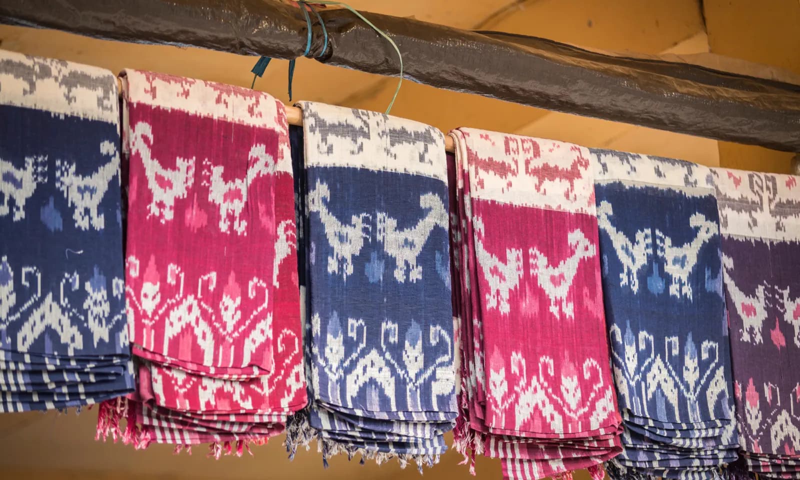 Traditional woven textiles with bird and floral motifs in blue, pink and white hanging folded over a wooden beam