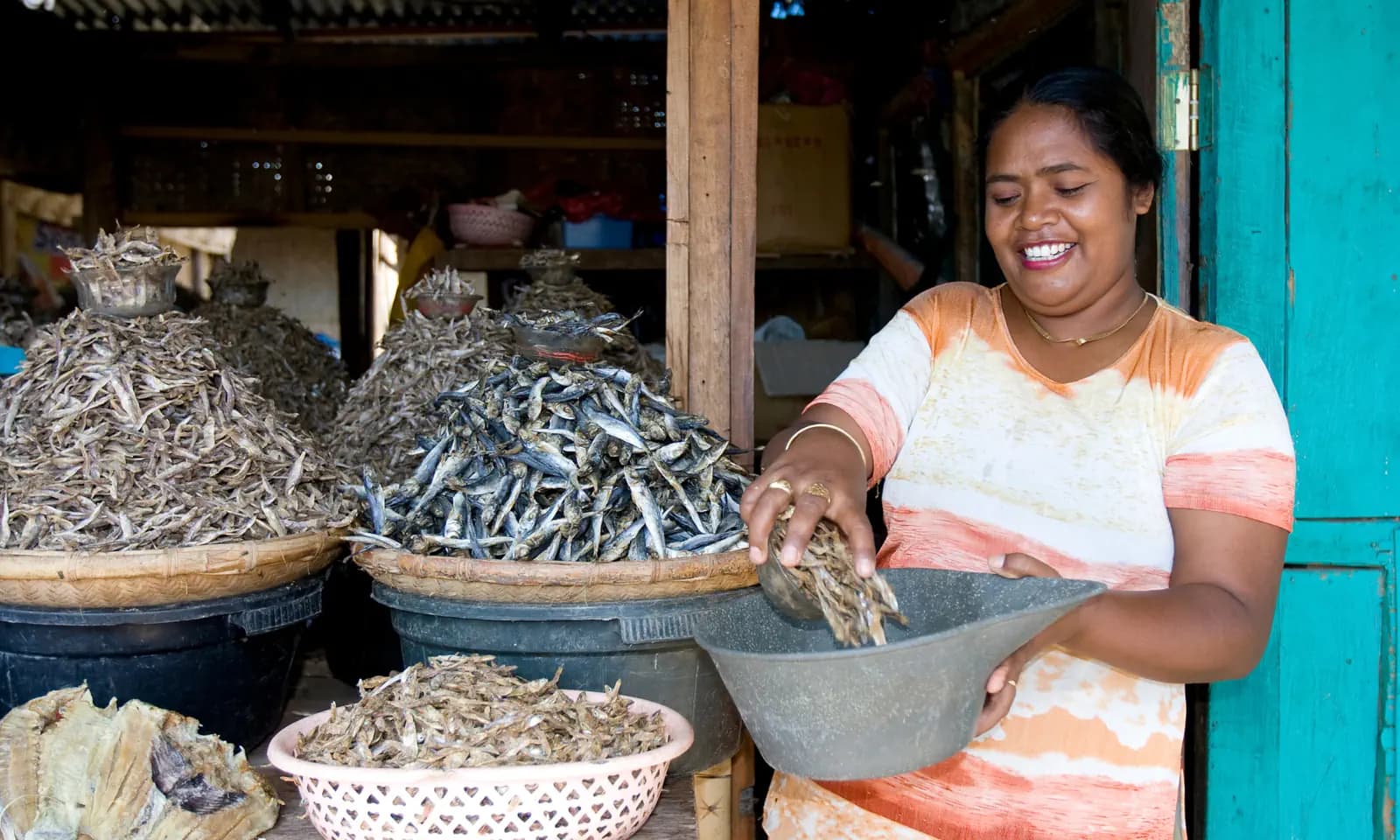 Woman smiling while scooping dried fish from a large basket at a market stall