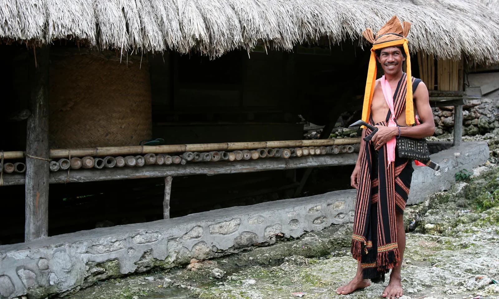 Man in traditional woven headdress and sarong standing barefoot before a thatched-roof house with stone-walled foundation