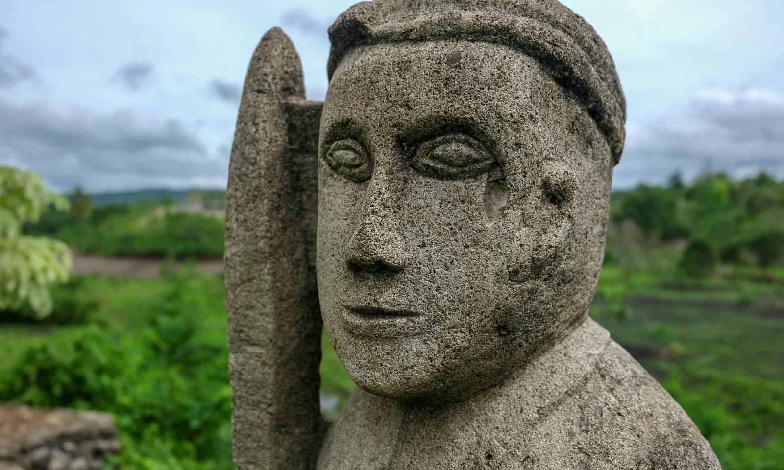 Close-up of a weathered stone statue's face with a carved headpiece, greenery and cloudy sky in the background