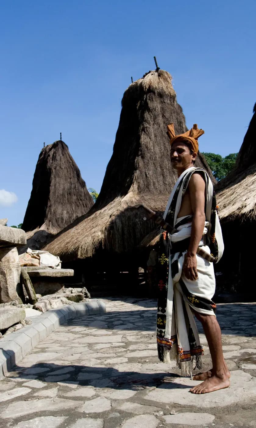 Man in traditional woven cloth and headband standing before tall thatched-roof houses with peaked grass roofs