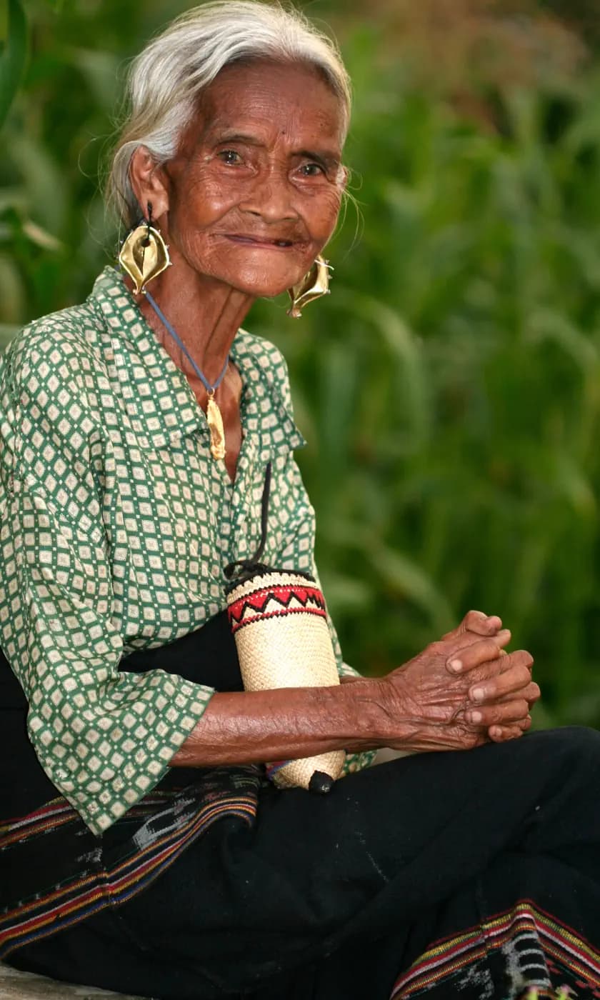 Elderly woman with gray hair and large gold earrings sitting outdoors, wearing a green patterned blouse and woven armband