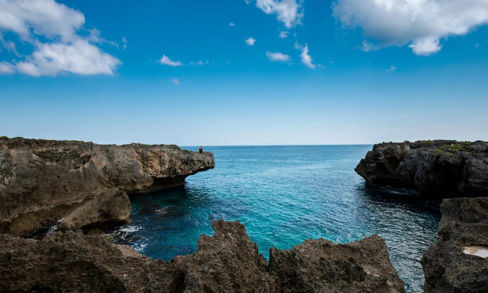 Rocky limestone cliffs frame a turquoise inlet, with a person standing at the edge of the far outcrop