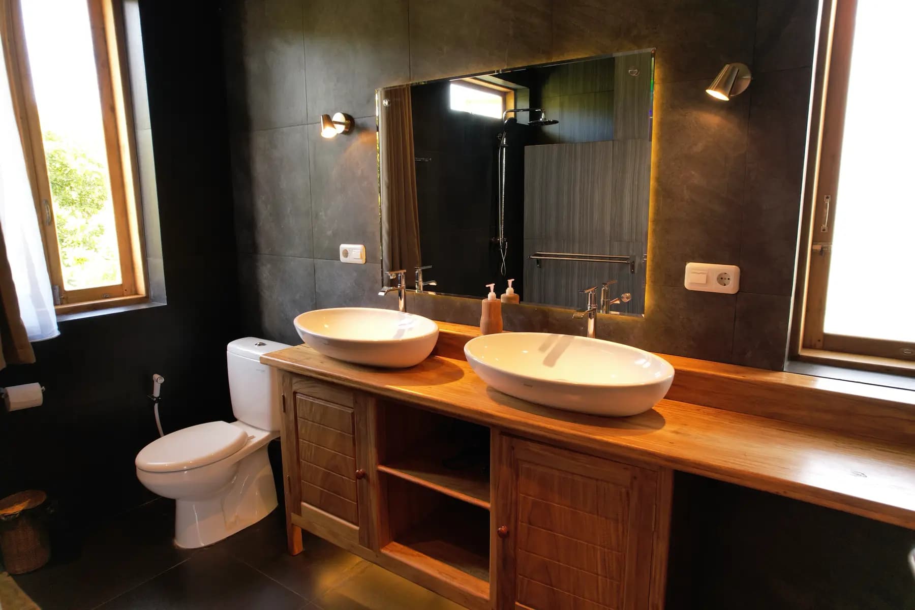 Bathroom at Akasha Beach with double white vessel sinks on wooden vanity, large mirror, dark tile walls, toilet, and windows with natural light