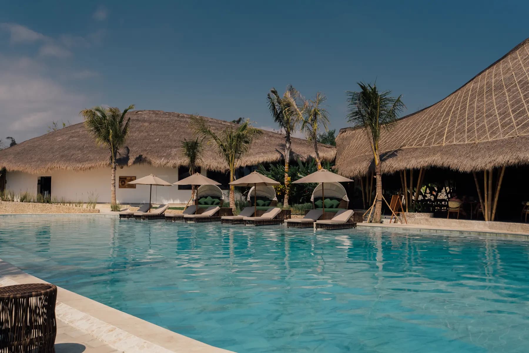 ARYA Sumba Resort pool area with turquoise water, thatched-roof buildings, palm trees, and lounge seating with umbrellas