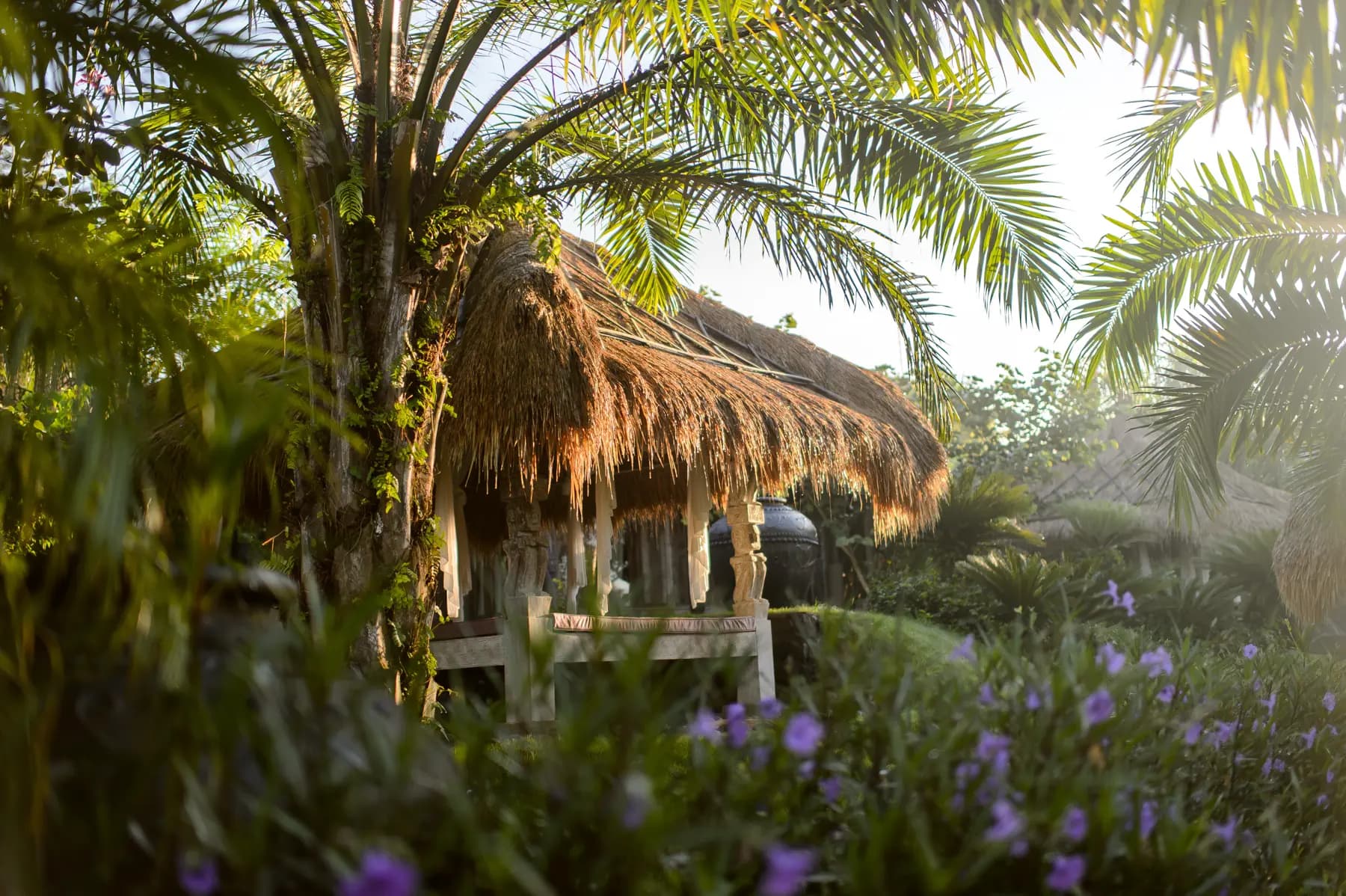 Thatched-roof bungalow surrounded by tropical vegetation and purple flowers, with palm trees overhead at Blue Karma Djiwa Ubud