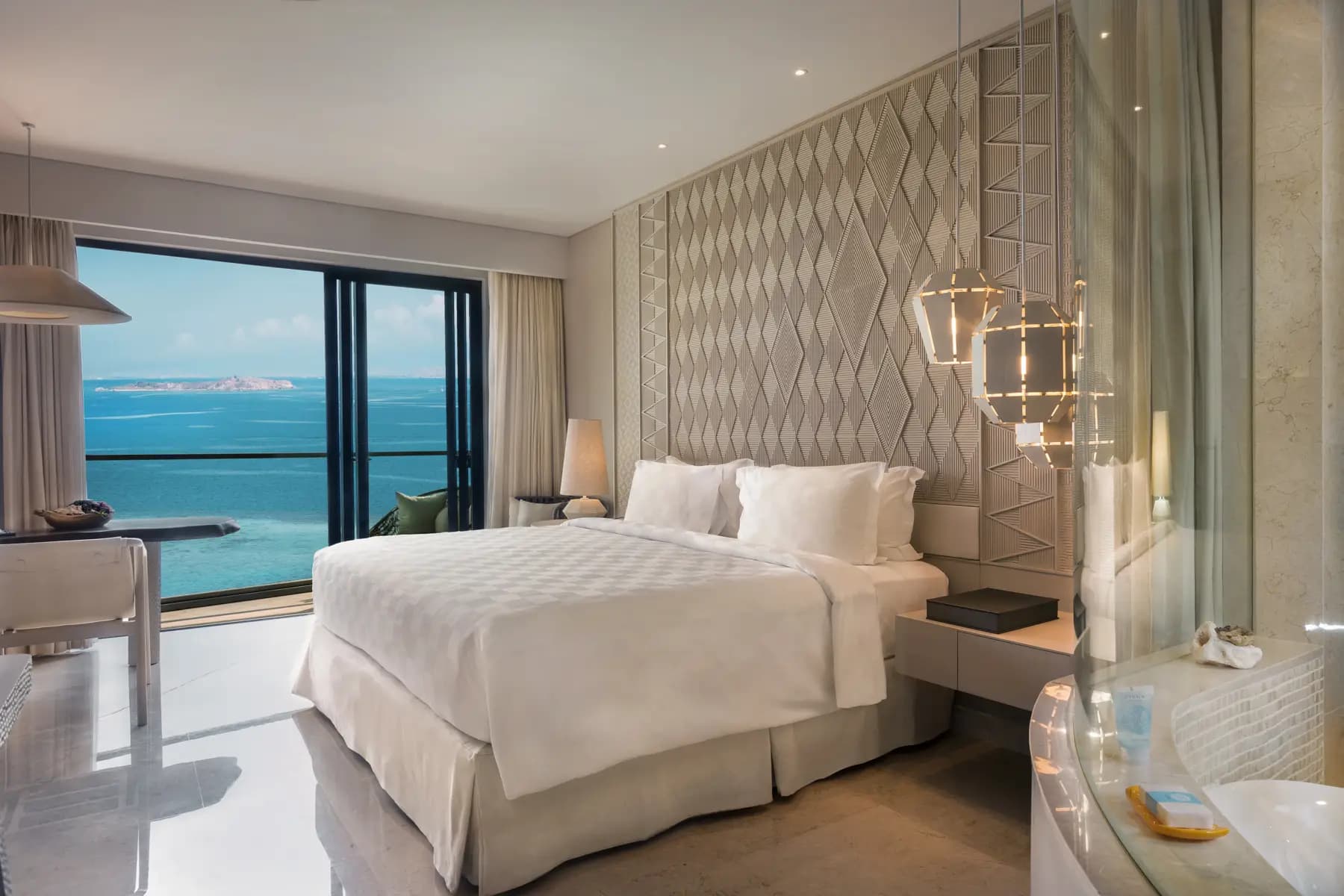 Bedroom with queen bed, textured headboard, pendant lighting, bedside tables, and floor-to-ceiling windows with a blue water view
