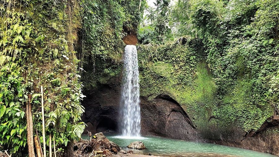 A narrow waterfall drops through a rock overhang into a turquoise pool surrounded by dense green cliffside foliage