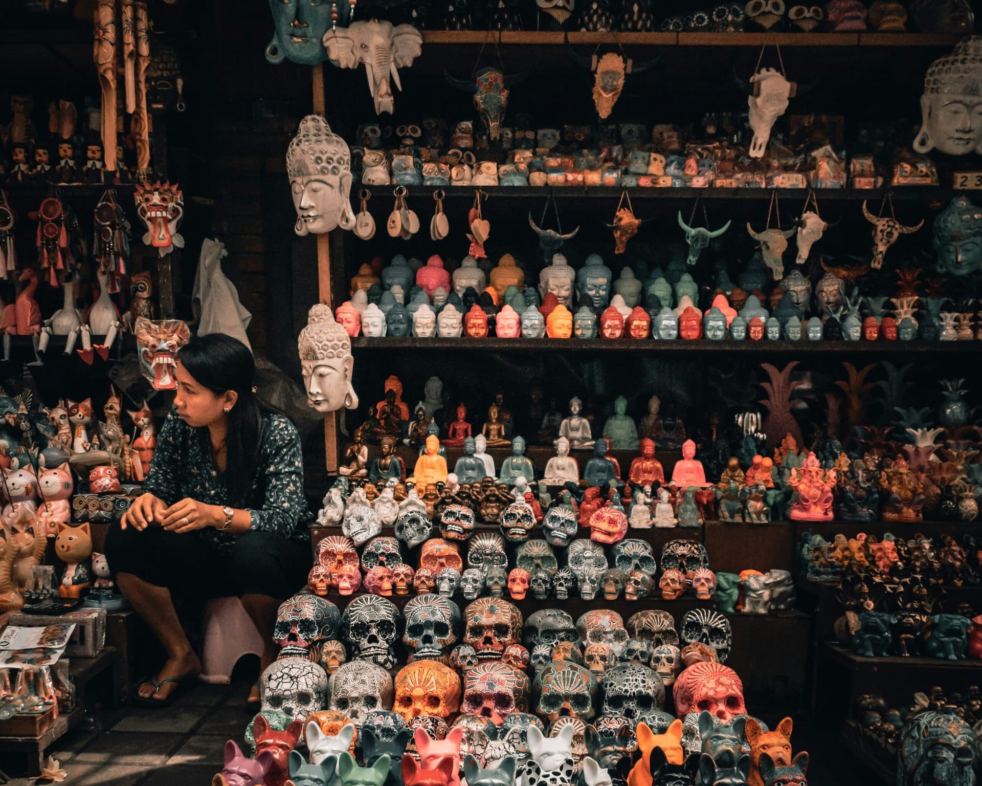 A vendor sits among rows of carved wooden masks, painted skull figurines, Buddha heads and animal carvings on a market stall