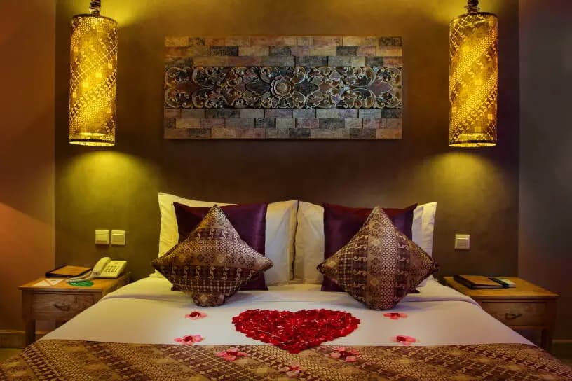 A bedroom at Mathis Retreat Ubud with a bed decorated with red rose petals in a heart shape, patterned cushions, dark walls, pendant lights, and artwork above the headboard