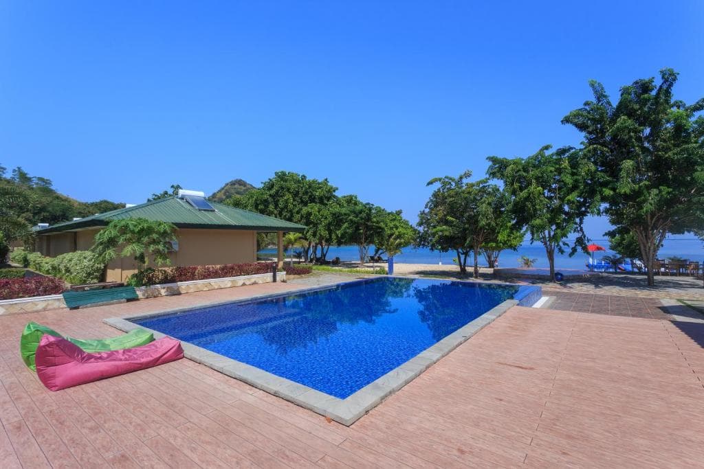 Sylvia Hotel & Resort Komodo's swimming pool with brick paving, pink and green inflatable seating, a building with green roof, mature trees, and water visible beyond
