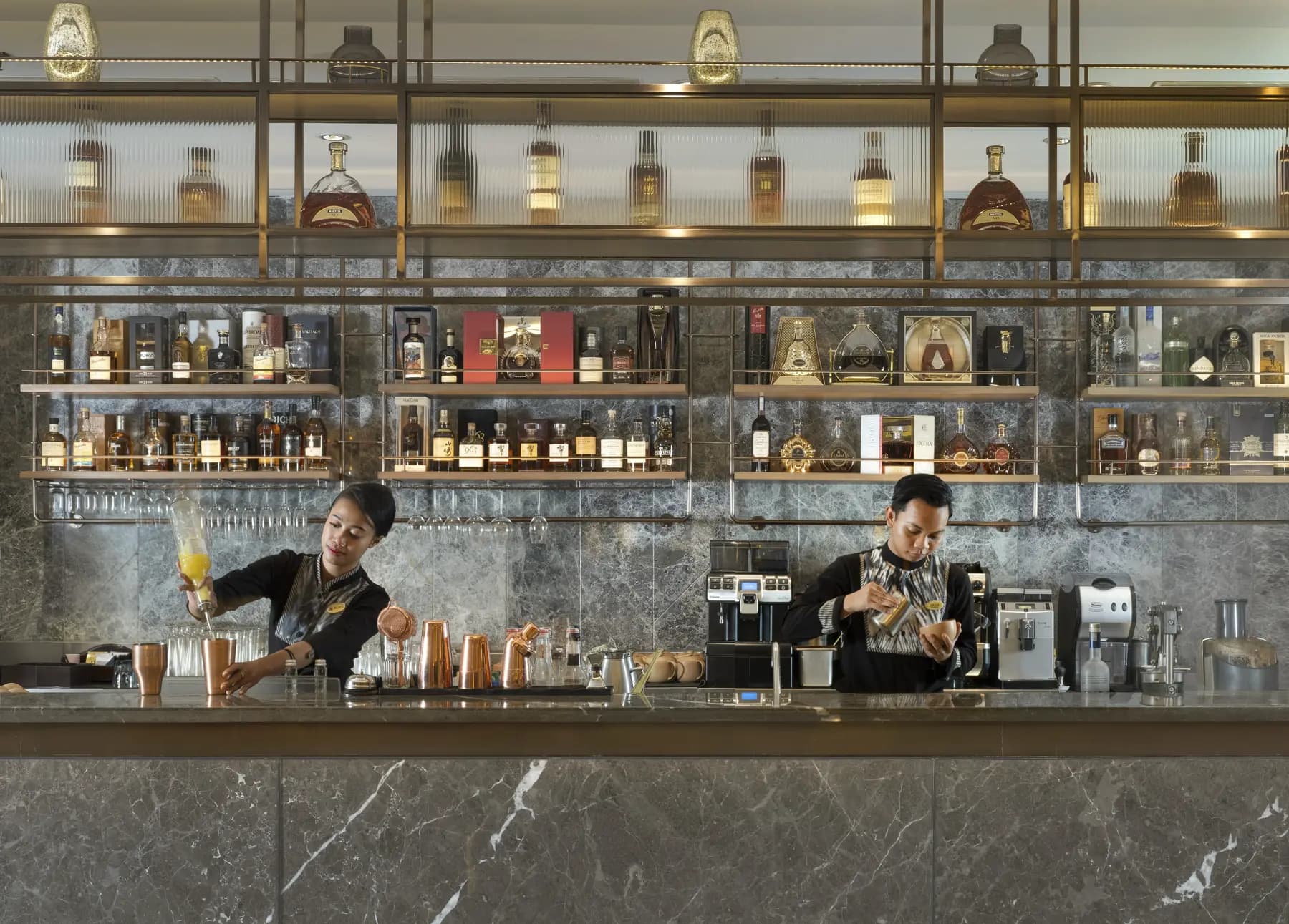 Two bartenders work behind a counter at Royal Ambarrukmo's bar, with shelves of bottles and glassware displayed on the wall behind them
