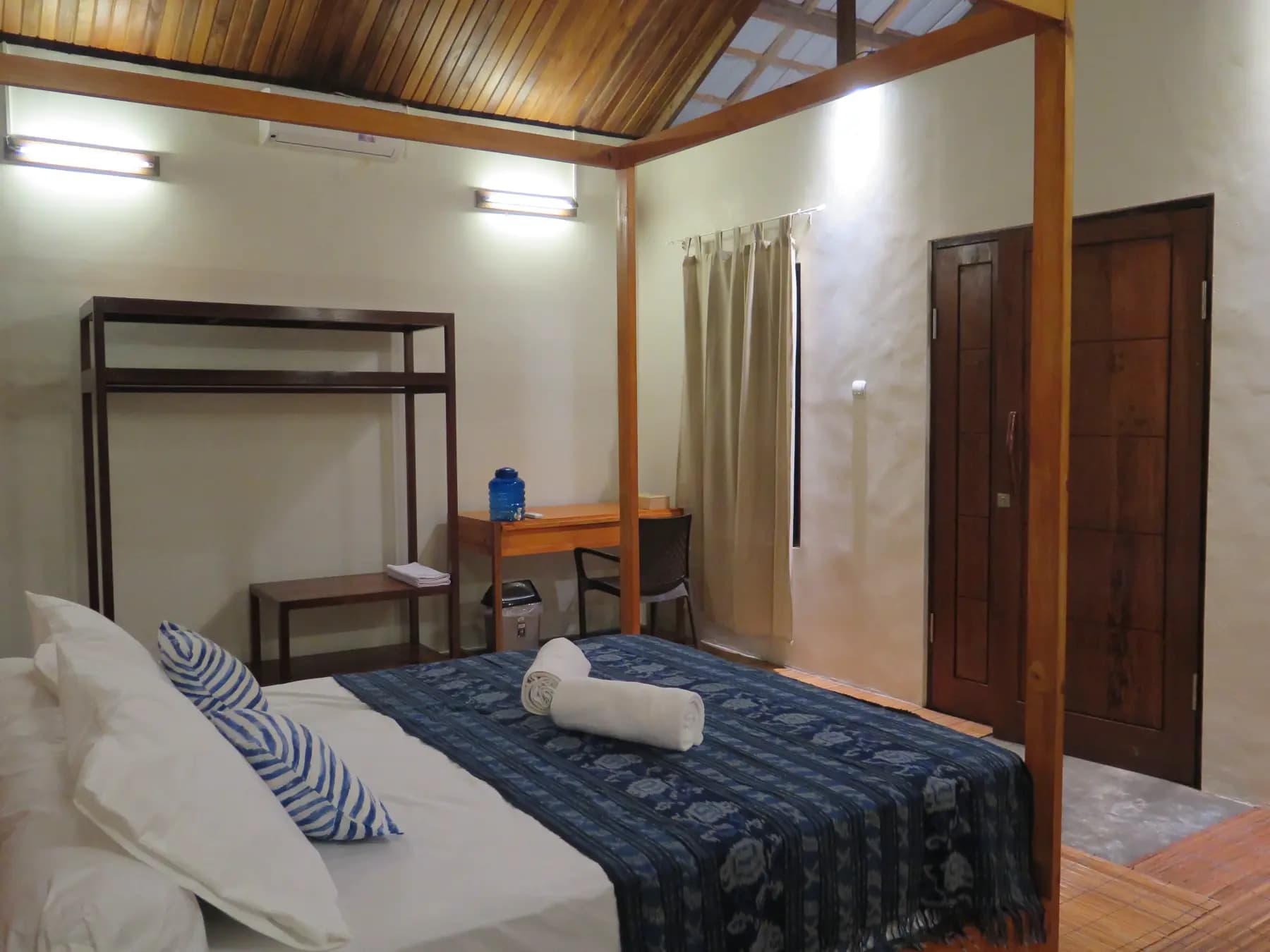 HamuEco Dive Resort bedroom with wooden four-poster bed, desk, chair, two wooden doors, and wooden ceiling beams