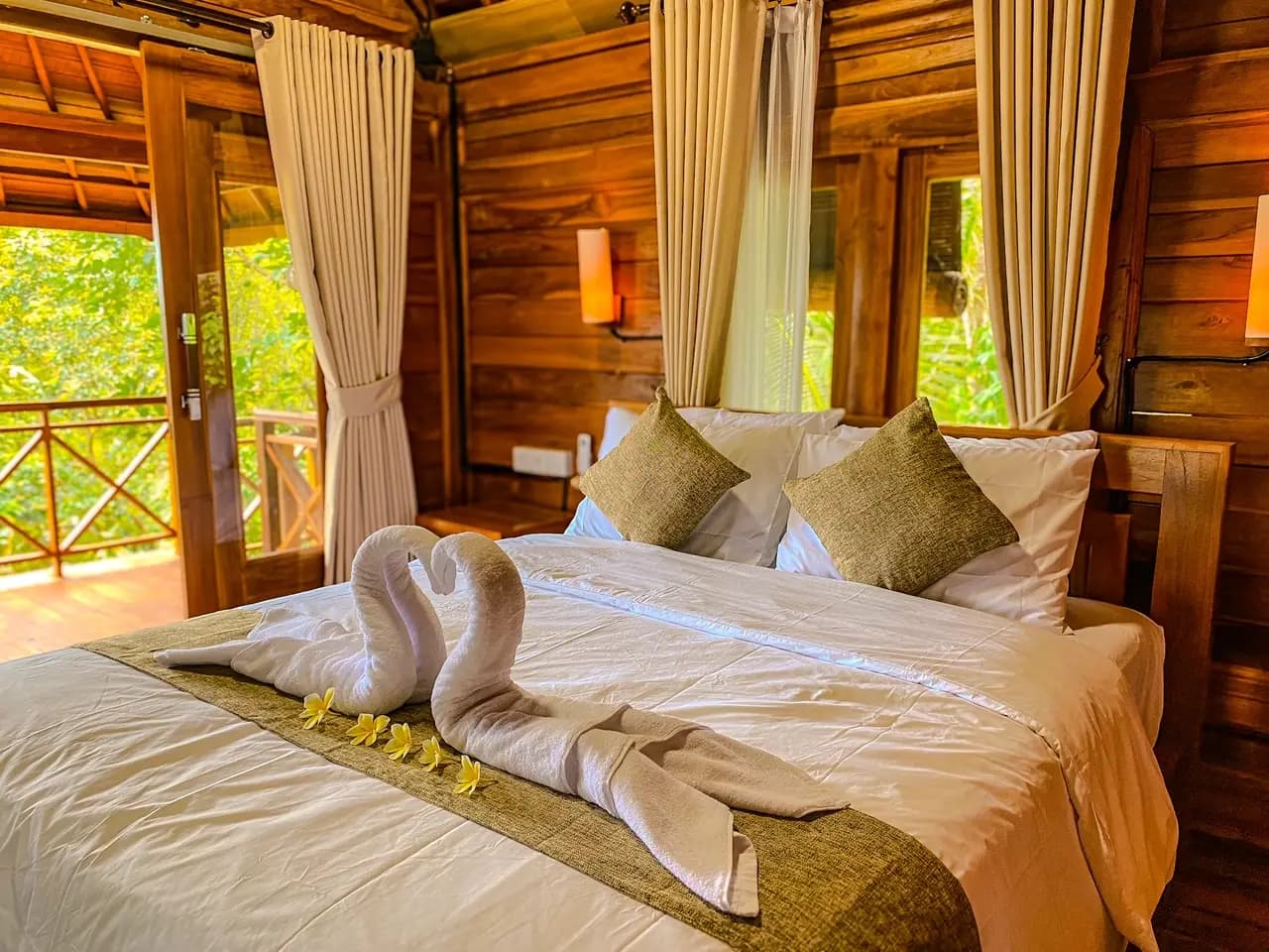 Bedroom at Wimala Farm House with a bed decorated with towel swans, green cushions, white bedding, wooden walls, curtained doors opening onto a balcony, and green vegetation visible outside