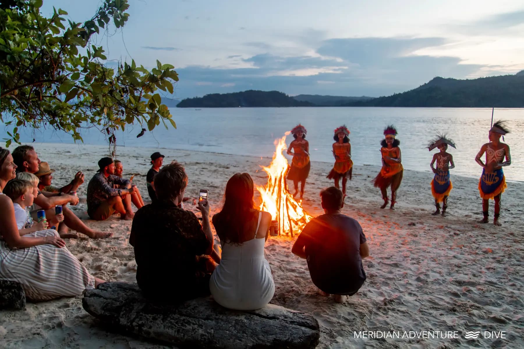 Guests and local people gathered around a bonfire on a beach at dusk, with water and forested shoreline visible beyond