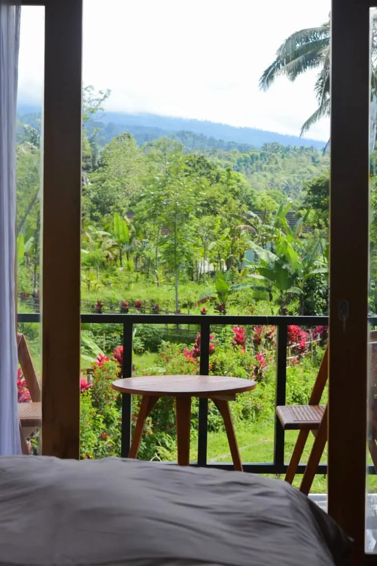 View from a bedroom at Mimpi Manis Villa showing a small table and chairs on a covered balcony overlooking tropical vegetation and distant forested landscape