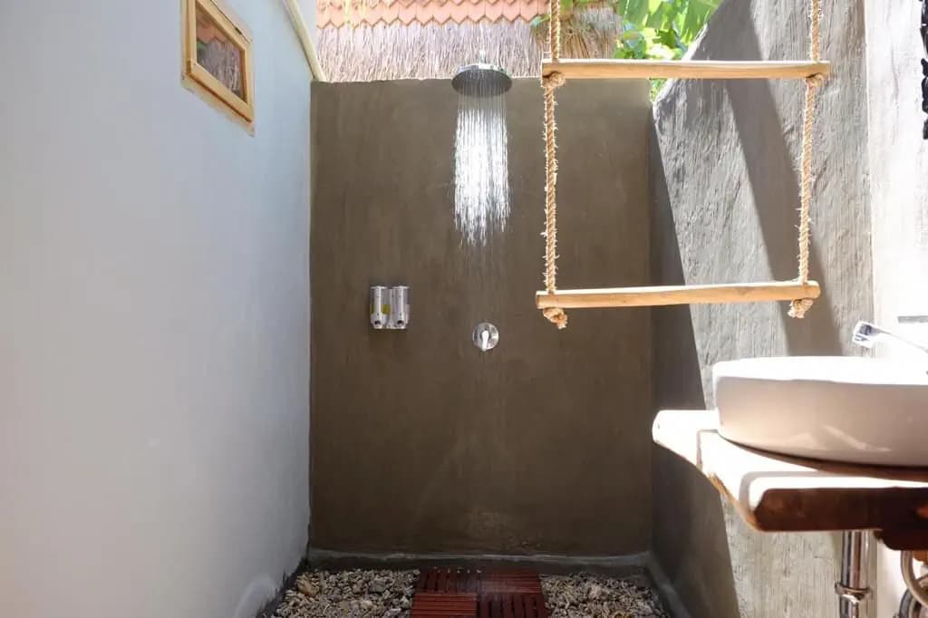 Outdoor shower at Tentacles Bali Cottage with dark tile walls, rainfall showerhead, wooden towel rack, and stone flooring