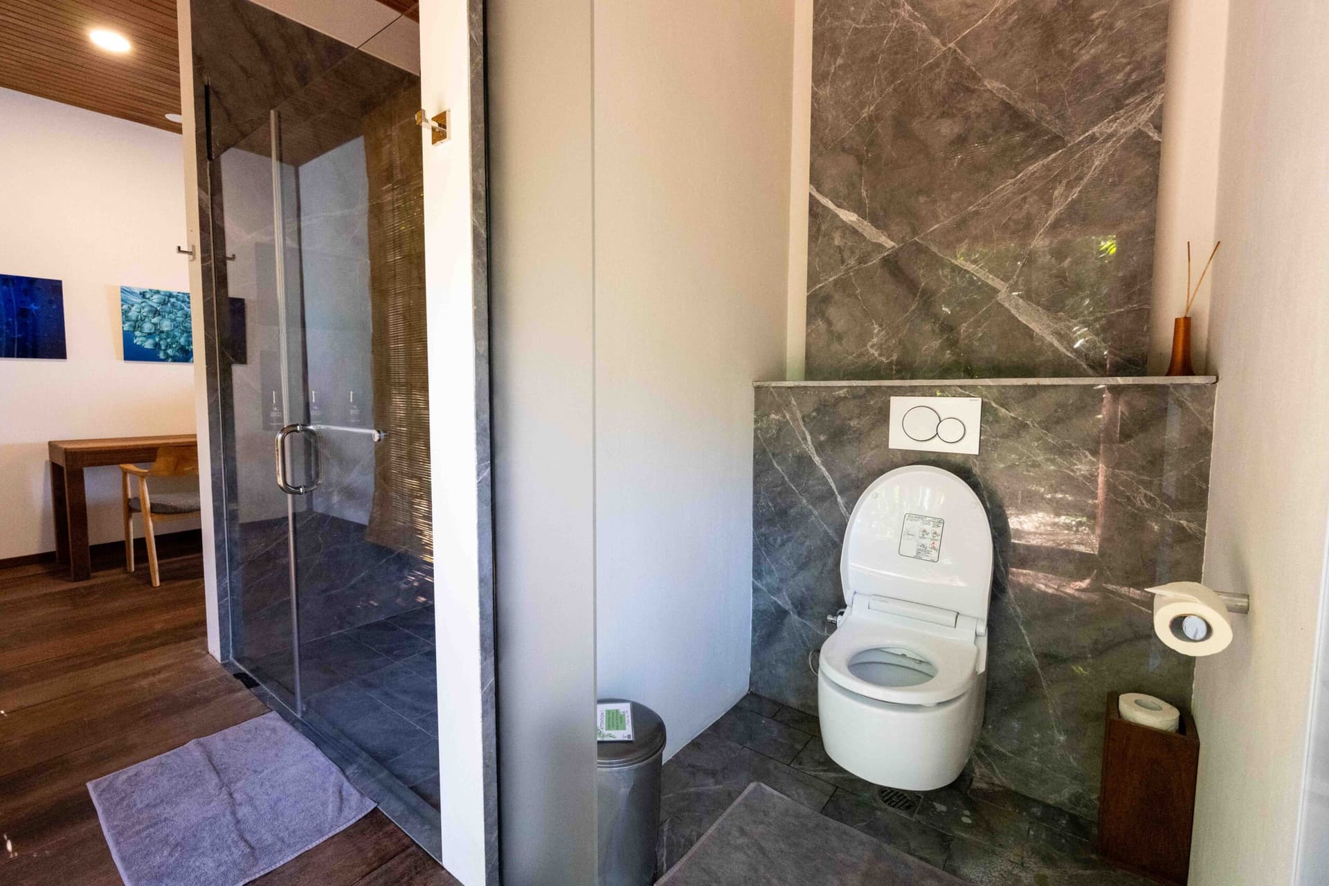 Modern bathroom at Sorido Bay Resort with dark marble walls, white toilet, glass shower enclosure, and wooden flooring visible in adjoining areas