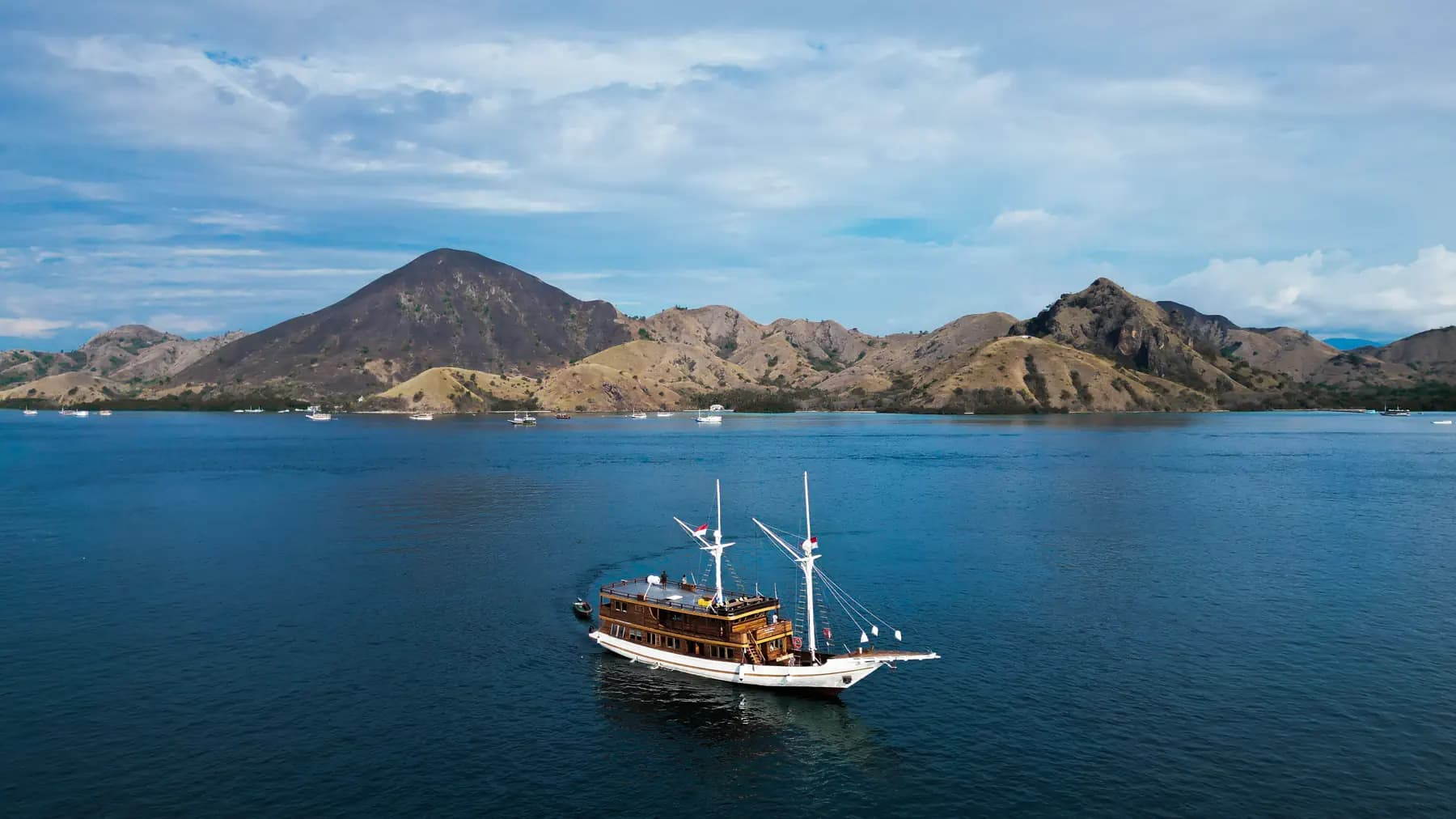 Sundari Voyages liveaboard with two masts anchored in calm blue water near rugged hills under a partly cloudy sky