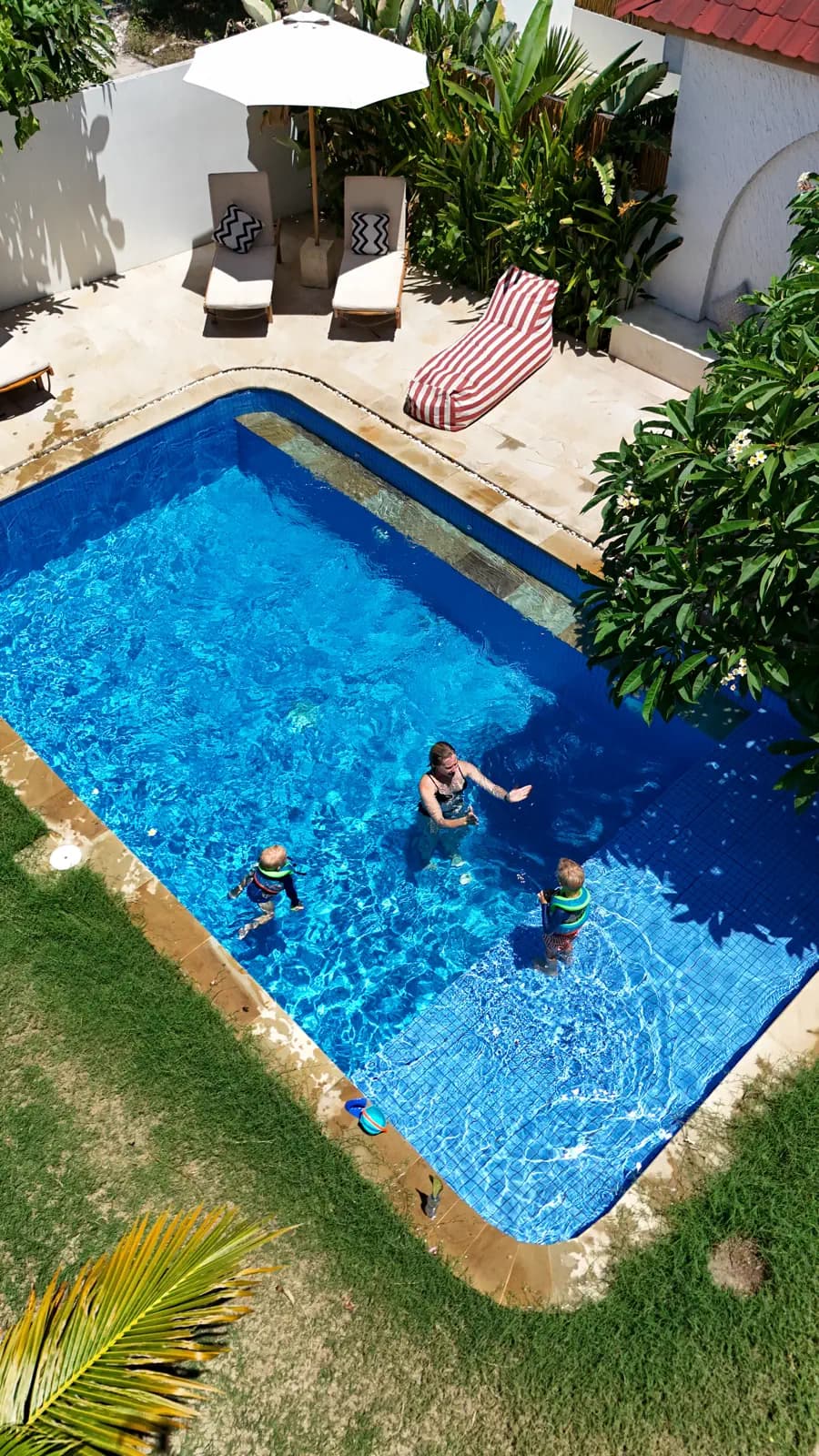 Villa Mila Gili Air's rectangular swimming pool with three guests, surrounded by sun loungers, parasol, potted plants, and grassed garden area