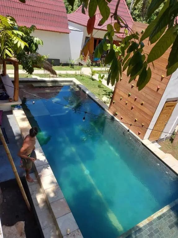 Twiny's lap pool surrounded by wooden deck, white walls, and tropical vegetation under a pink-roofed structure, with a guest in the water