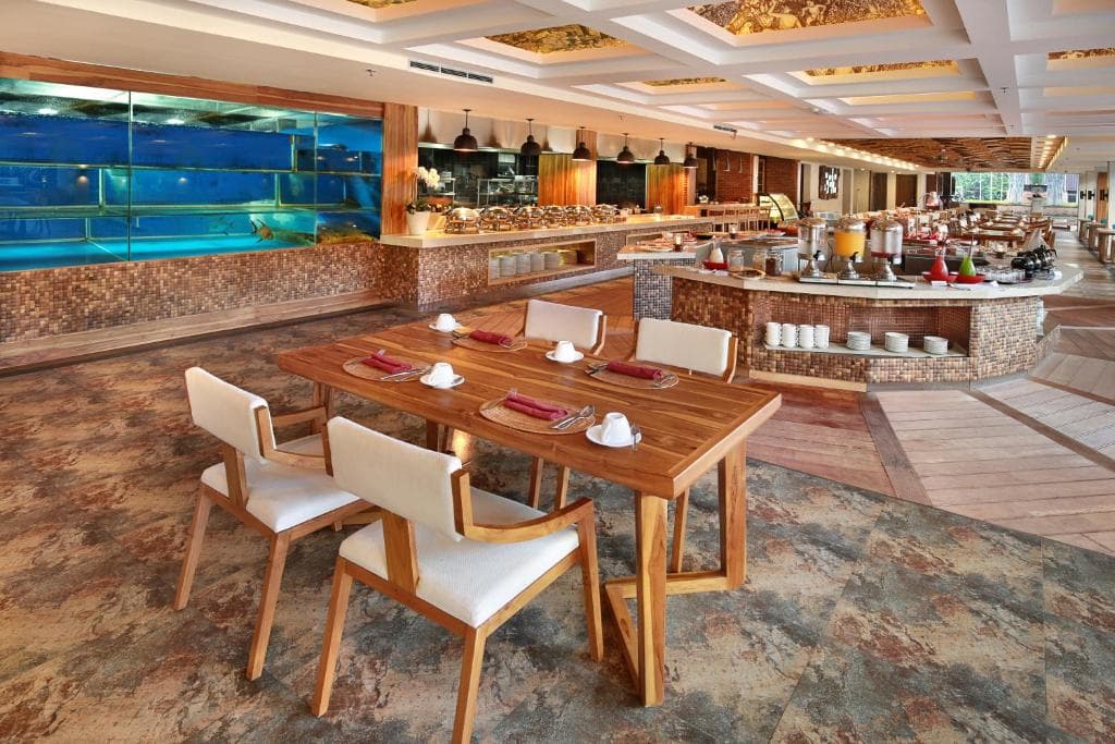 Jimbaran Bay Beach Resort & Spa restaurant interior with wooden dining table, chairs, service counter, and large windows overlooking water