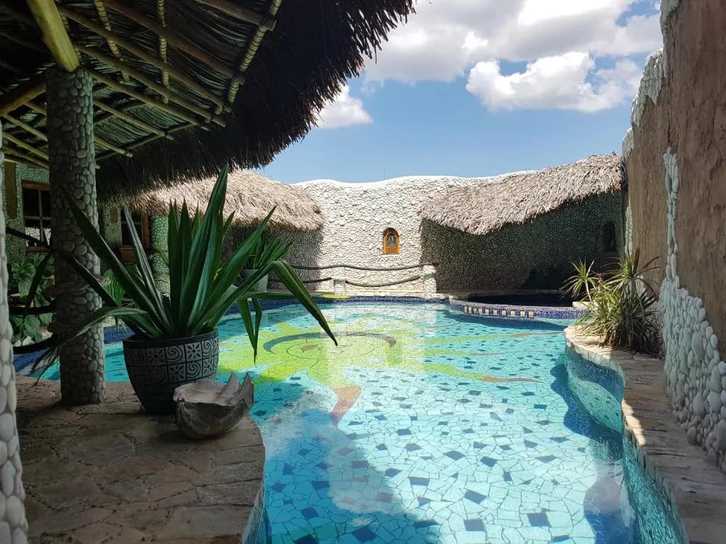 La Hasienda's pool courtyard features turquoise mosaic tiles, thatched roof structures, potted palms, and stone walls under blue sky