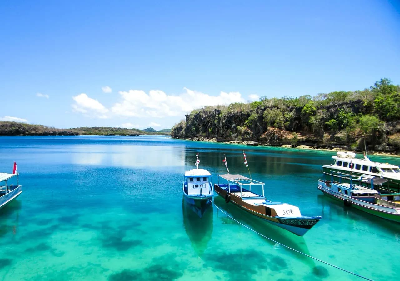 Wooden boats anchored in clear turquoise water beside a forested cliff under a blue sky with scattered clouds