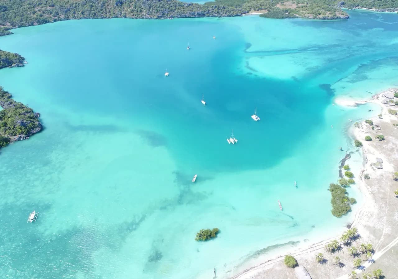 Aerial view of a turquoise lagoon with several small boats anchored near a sandy, palm-fringed shoreline