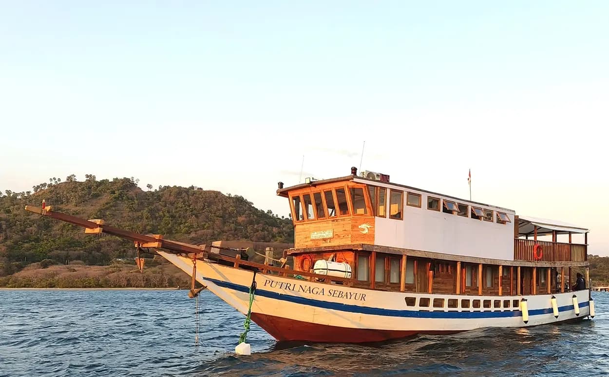 Putri Naga liveaboard boat anchored on calm water with a wooded hillside in the background at dusk