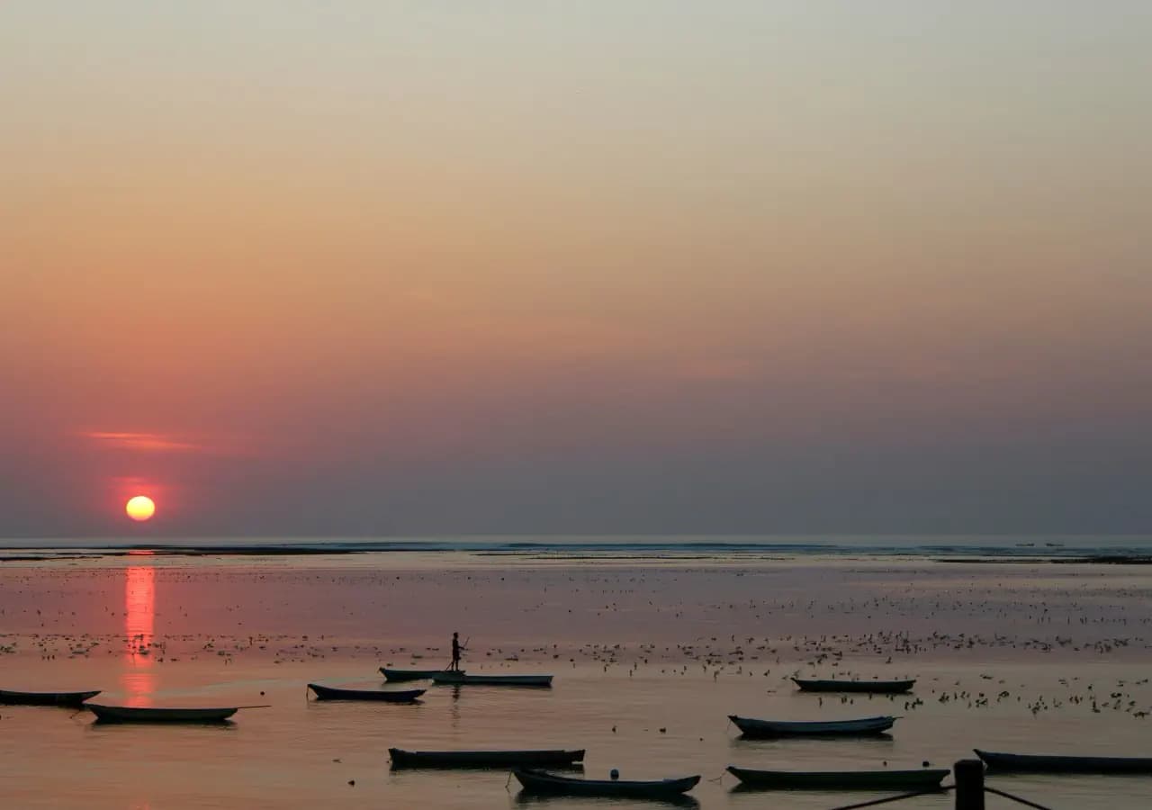 Silhouetted fishing boats and a person standing in shallow water at sunset, with the sun low over a calm sea