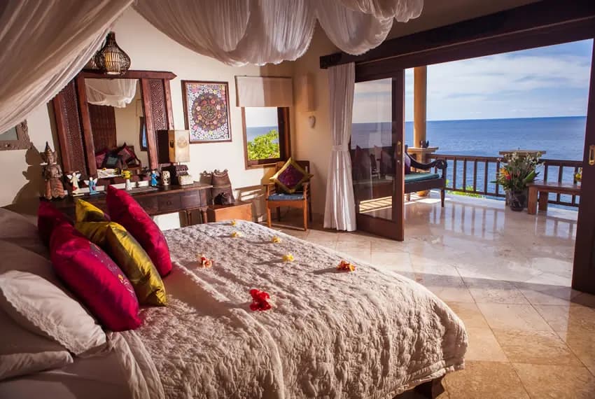 Spacious bedroom with draped canopy, colorful pillows on bed, vanity mirror, decorative items, and open doors leading to a balcony overlooking water