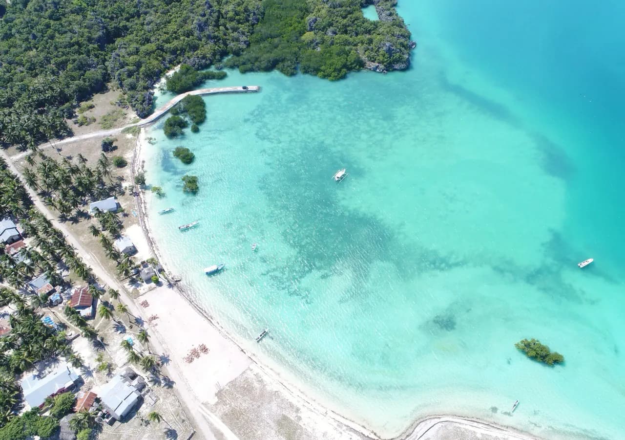 Aerial view of a curved white-sand beach and turquoise lagoon with small boats, bordered by dense trees and a wooden jetty
